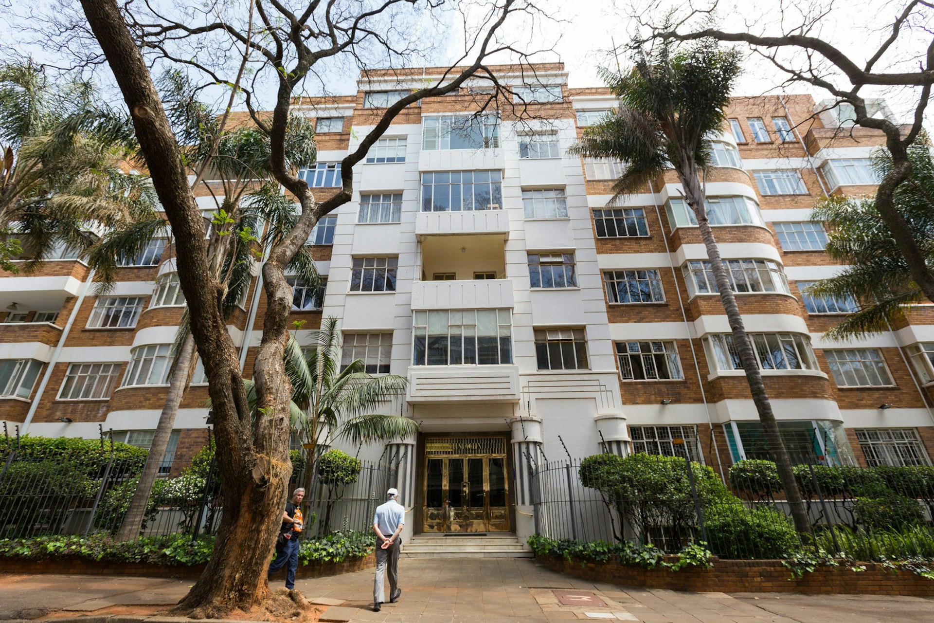 A elderly guide walks towards the golden doors of a towering art deco block of flats on one of the Johannesburg walking tours put on by the Heritage Foundation © Heather Mason / Lonely Planet