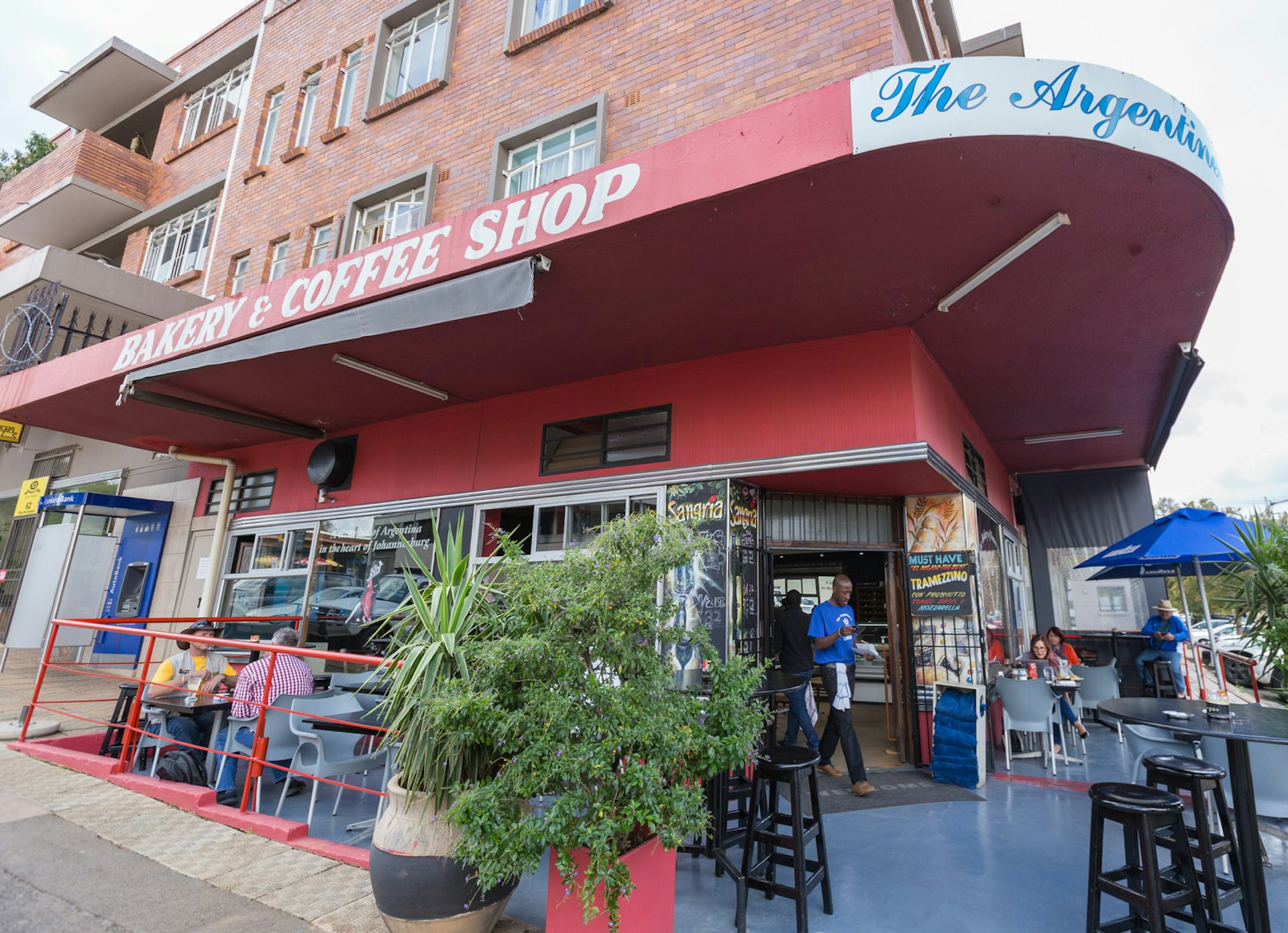 At the base of a large brick building is a bakery and coffee shop. Sitting on the corner, it has tables running down both streets. Patrons sit under umbrellas and dine, while a waiter walks out from the interior. Linden is one of Johannesburg's best neighbourhoods © Heather Mason / Lonely Planet
