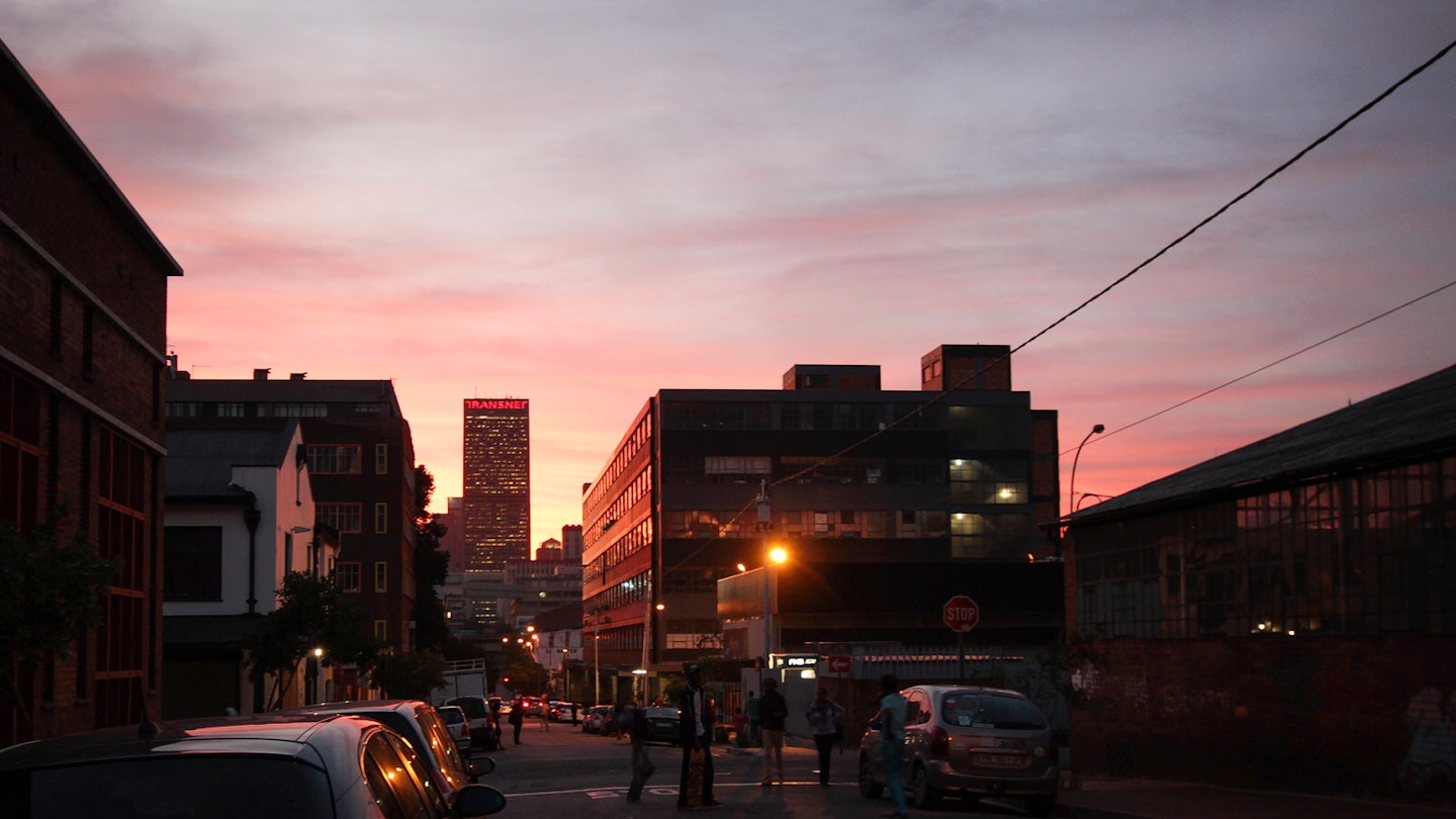 A shot of an intersection, with a bright pink-and-orange sky backing the surrounding buildings and traffic © Heather Mason / Lonely Planet