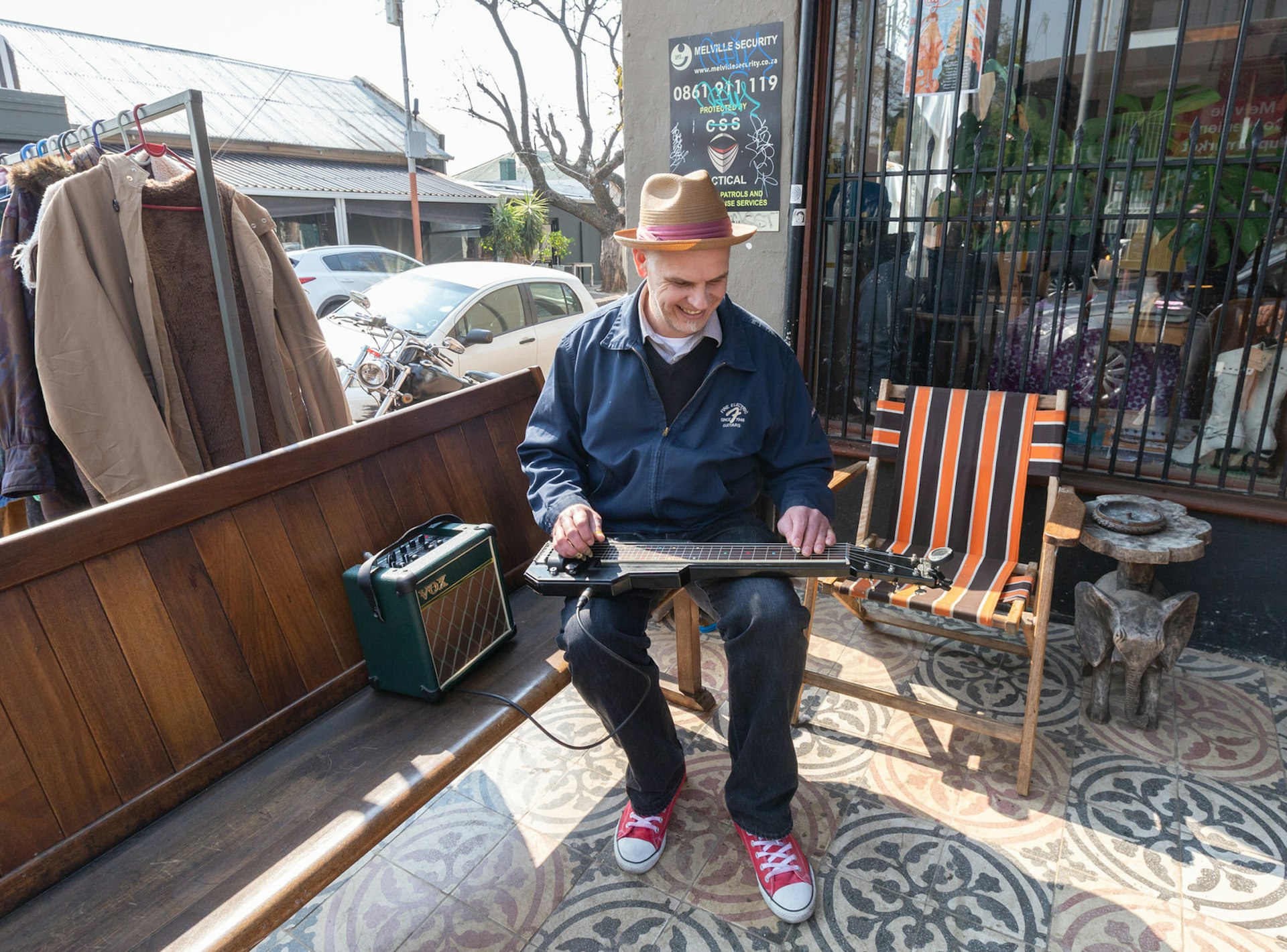 A hip-looking gentleman – in red converse Chuck Taylor shoes, a trilby hat and jeans – sits in a deck chair on the pavement and plays a guitar that is laid flat across his lap. Melville is one of Johannesburg's best neighbourhoods © Heather Mason / Lonely Planet