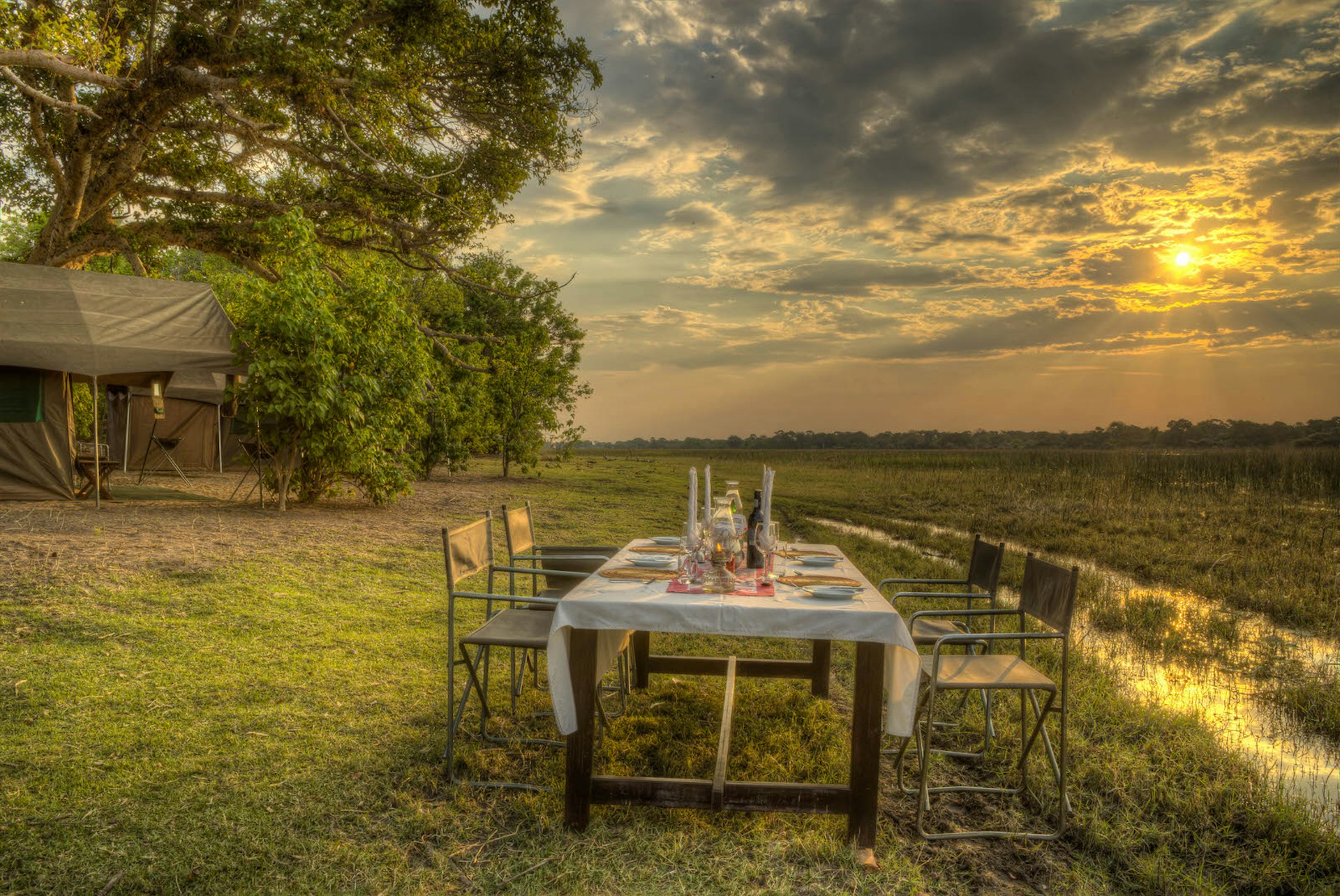 A wooden dining table, covered with a white tablecloth and laid with placemats, plates, candles and cutlery, stands next to a stream of water in an open savannah wilderness. To the left is a mobile tent beneath a towering tree © James Gifford / Lonely Planet