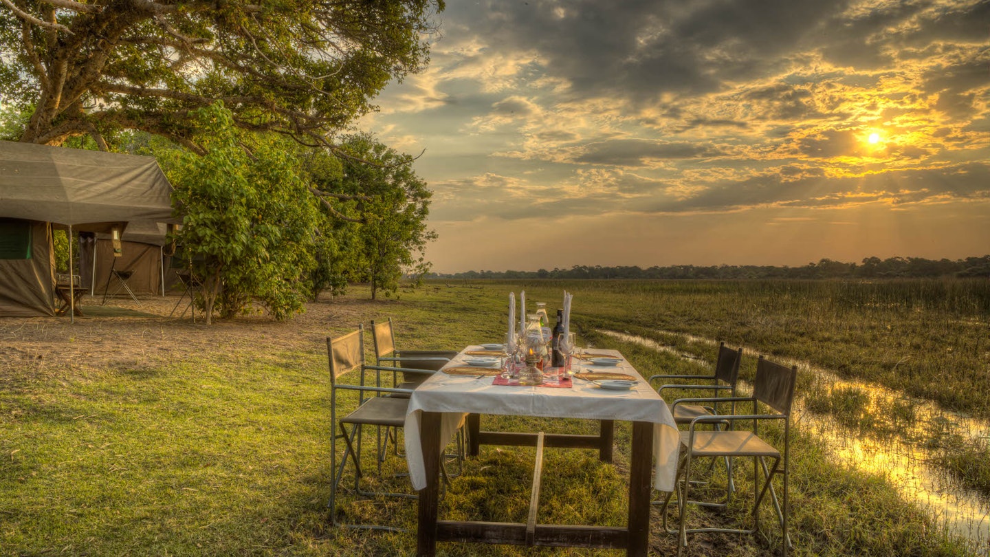 A wodden dining table, covered with a white table cloth and laid with place mats, plates, candles and cutlery, stands next to a stream of water in an open savannah winderness. To the left is a mobile tent beneath a towering tree © James Gifford / Lonely Planet