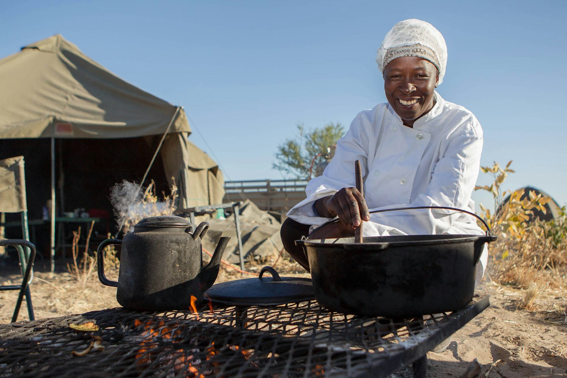 A smiling Botswanan chef, dressed in whites, crouches over a crockpot cooking over a fire in a mobile safari camp, with the portable tents seen in the background © James Gifford / Lonely Planet