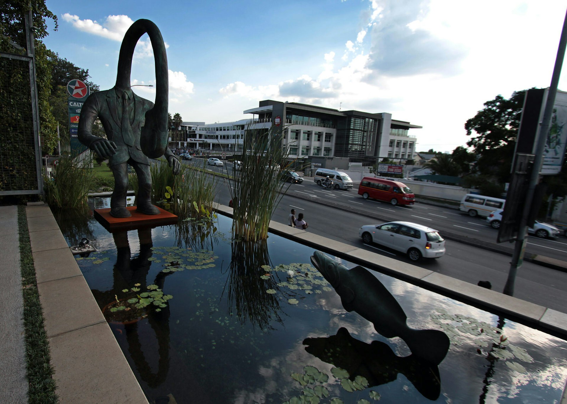 This image looks over a small rectangular-shaped pond that sits raised above the street's pavement. A statue of a fish jumps from one end of the pond, while a sculpture of a human with a long, elasticated neck stands at the other end. The human's neck arches over so that the human is looking back at its chest. Rosebank is one of Johannesburg's best neighbourhoods © Heather Mason / Lonely Planet