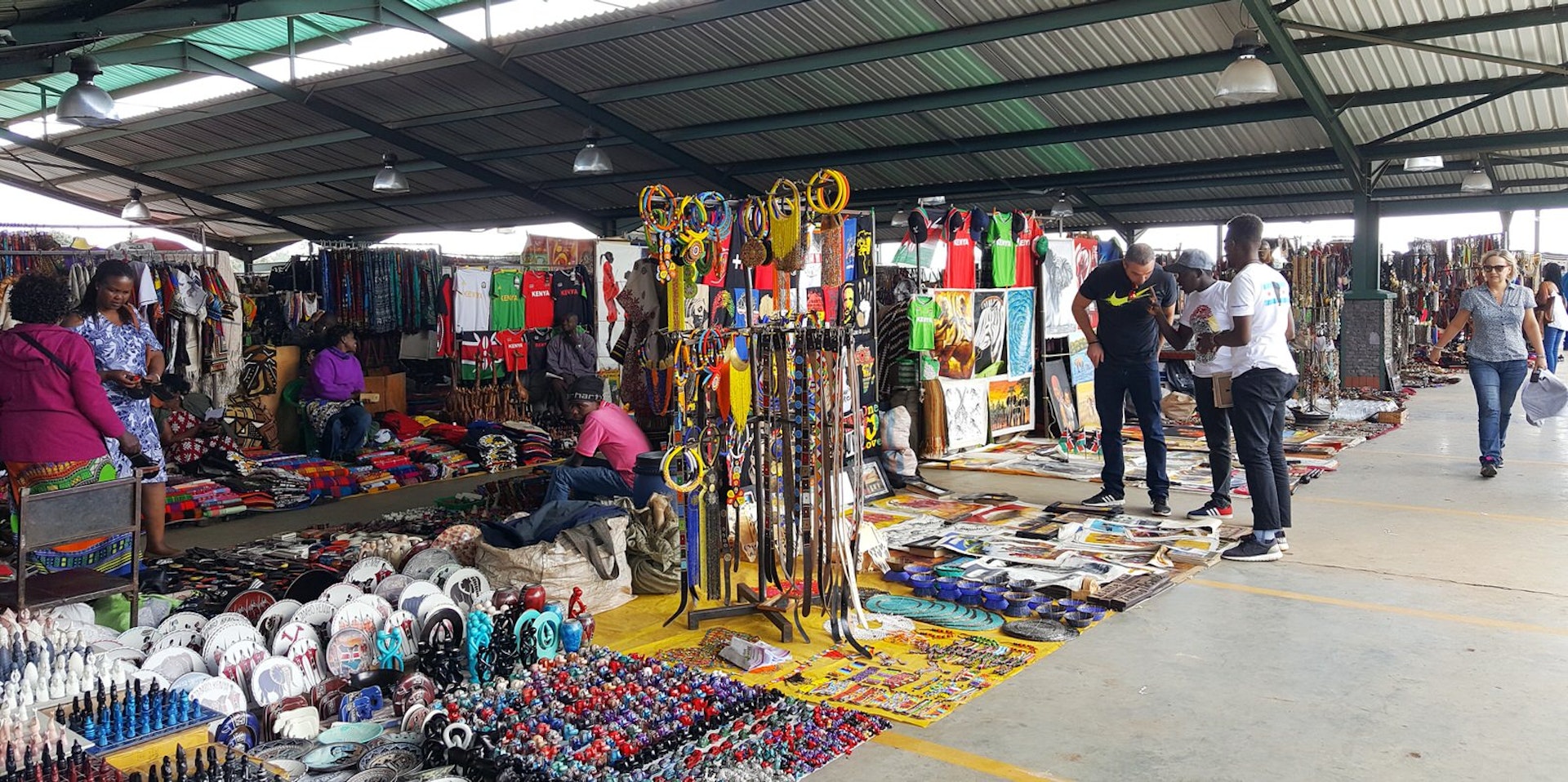 Under a modern, sloping roof of steel frames and corrugated tin cladding, is a bright market full of Maasai beaded necklaces, paitings, plates etc. Much is laid on the ground, other items sit on tables or hang from racks. There are a few tourists talking with vendors © Clementine Logan / Lonely Planet