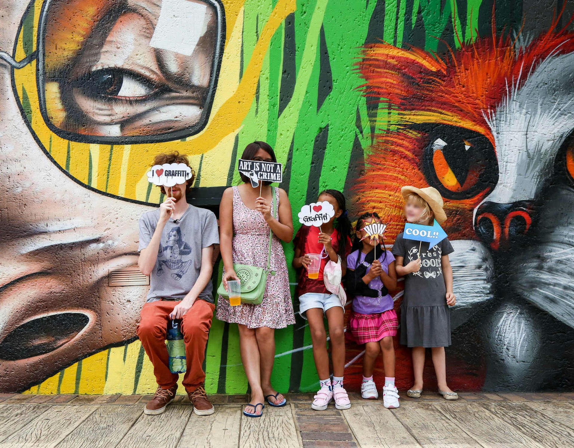 Two adults and three children stand in front of a wall covered in street art. The man holds a small cloud-shaped sign that covers his face and reads 'I heart graffiti'. The woman holds a similar sign that reads 'Art is not a crime'. The children hold similar sings, including ones that say 'Wow' and 'Cool'. They are on one of Past Experiences' Johannesburg walking tours © Heather Mason / Lonely Planet