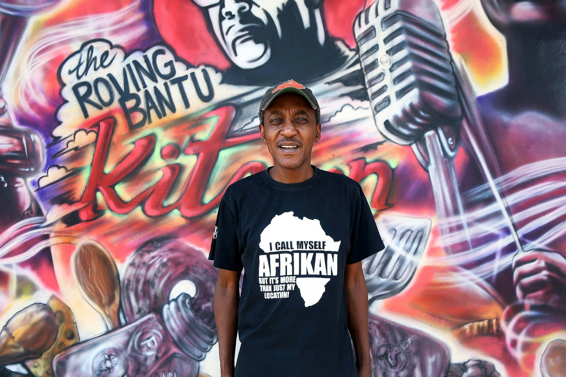 Sifiso Ntuli, stands in front of his restaurant which is covered in a street art mural. He wears a T-shirt that says, 'I all myself Afrikan, but it's more than just my location!' © Heather Mason / Lonely Planet