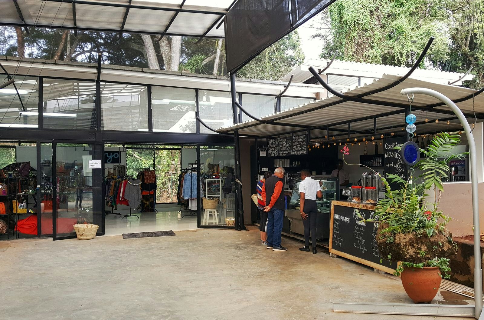 An elderly couple stand on a polished concrete floor in a bright, open-air atrium and ponder choices in a cafe display case. Above them is a view out to you lush trees and the sky. Inside the store beyond are various woven clothing items for sale © Clementine Logan / Lonely Planet