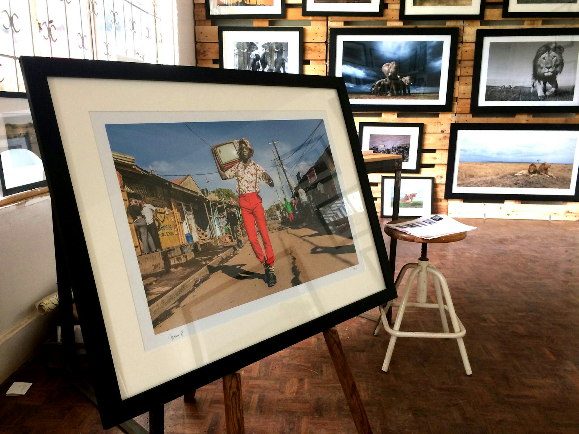 In the foreground is a large framed picture standing on an easel. In the picture stands a hip Kenyan man dressed in bright red trousers, flowered shirt, suspenders, sunglasses and sun hat. On his shoulders sits on old fashioned television. It looks to have been taken on the street in Kibera, Naiobi's largest slum. Behind this fram is a wall covered in framed photographs of Kenyan wildlife. The gallery is great for Nairobi shopping © Clementine Logan / Lonely Planet