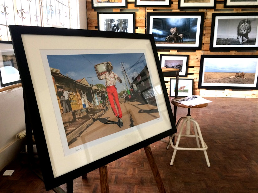 In the foreground is a large framed picture standing on an easel. In the picture stands a hip Kenyan man dressed in bright red trousers, flowered shirt, suspenders, sunglasses and sun hat. On his shoulders sits on old fashioned television. It looks to have been taken on the street in Kibera, Naiobi's largest slum. Behind this fram is a wall covered in framed photographs of Kenyan wildlife © Clementine Logan / Lonely Planet