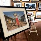 In the foreground is a large framed picture standing on an easel. In the picture stands a hip Kenyan man dressed in bright red trousers, flowered shirt, suspenders, sunglasses and sun hat. On his shoulders sits on old fashioned television. It looks to have been taken on the street in Kibera, Naiobi's largest slum. Behind this fram is a wall covered in framed photographs of Kenyan wildlife © Clementine Logan / Lonely Planet