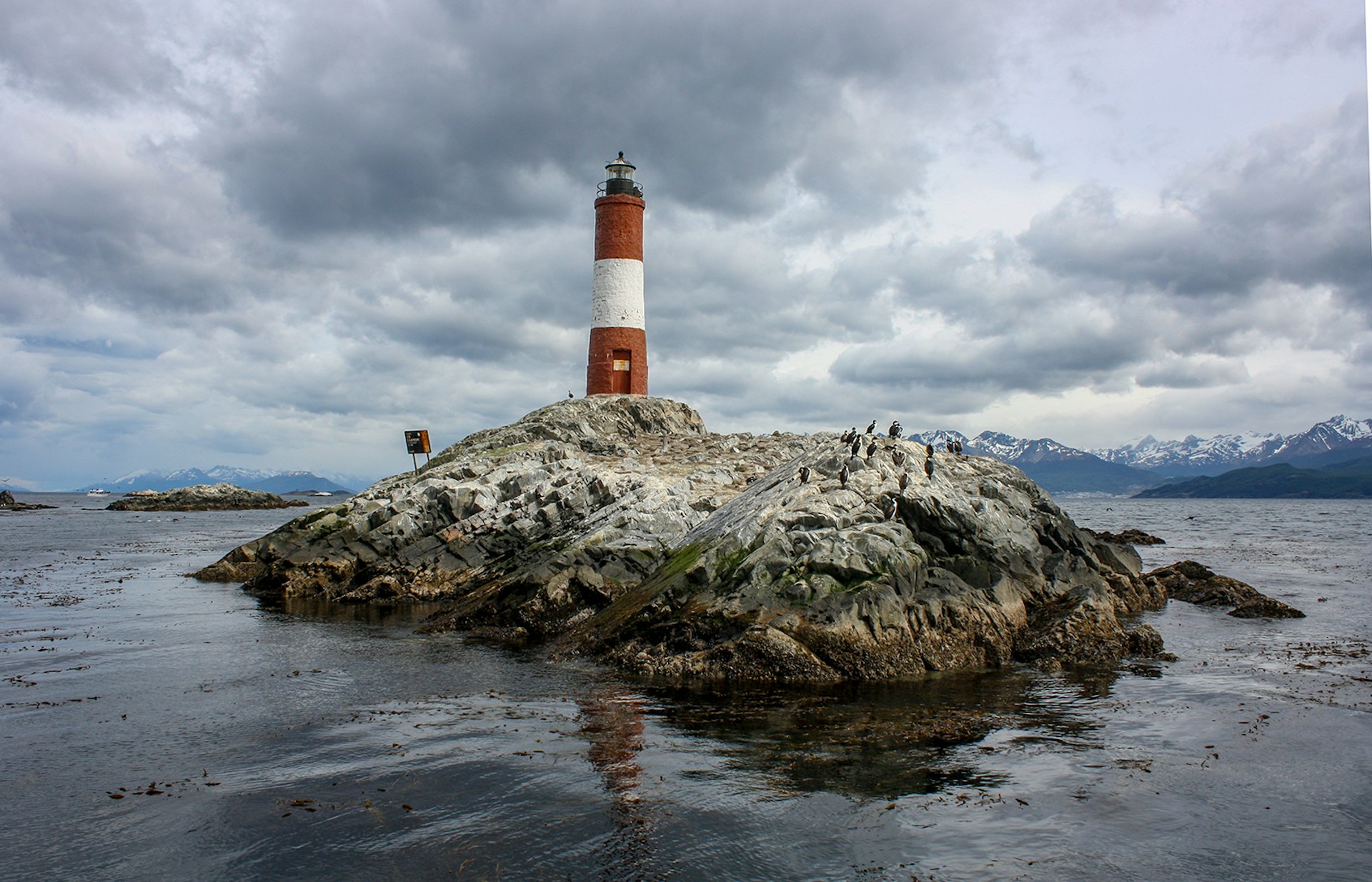 A red and white lighthouse on a rock, with penguins standing in the foreground and mountains framing the scene © Bridget Gleeson / Lonely Planet