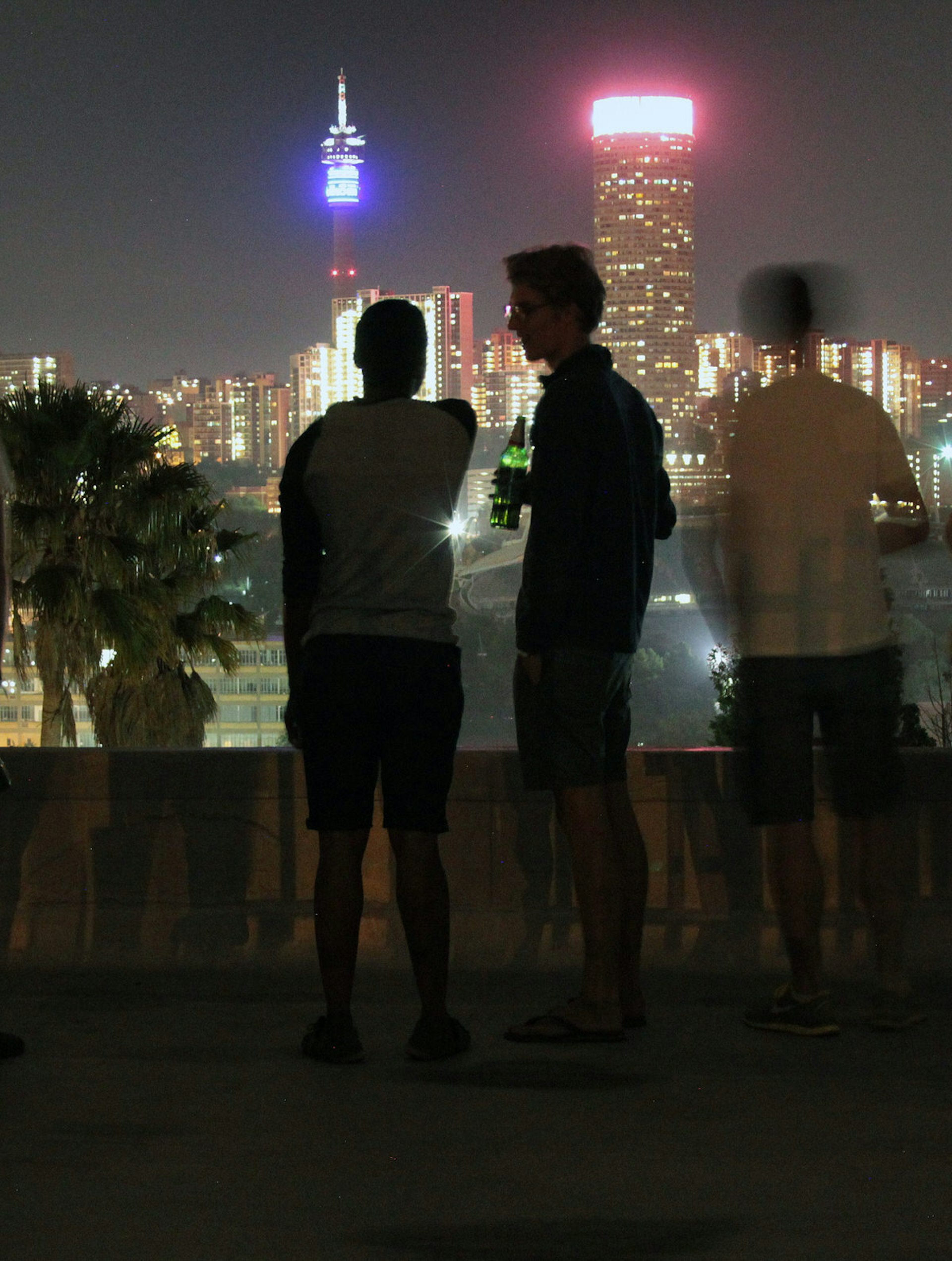A few people stand drinking on a dark rooftop and overlook a distant skyline of Joburg © Heather Mason / Lonely Planet