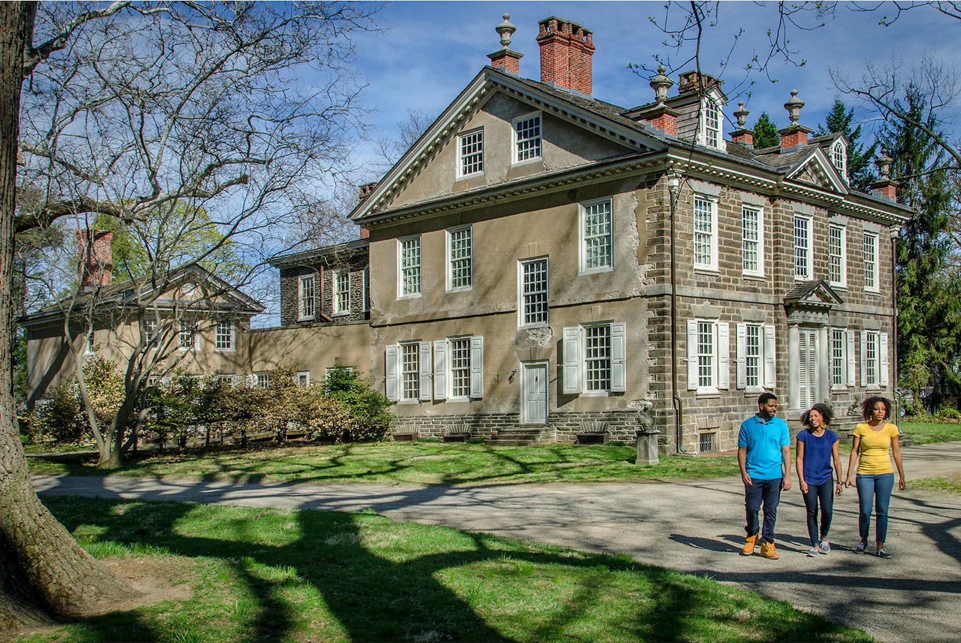 Three people walk outside of Cliveden, a three-story stone house built in 1767, on a sunny day © Visit Philadelphia