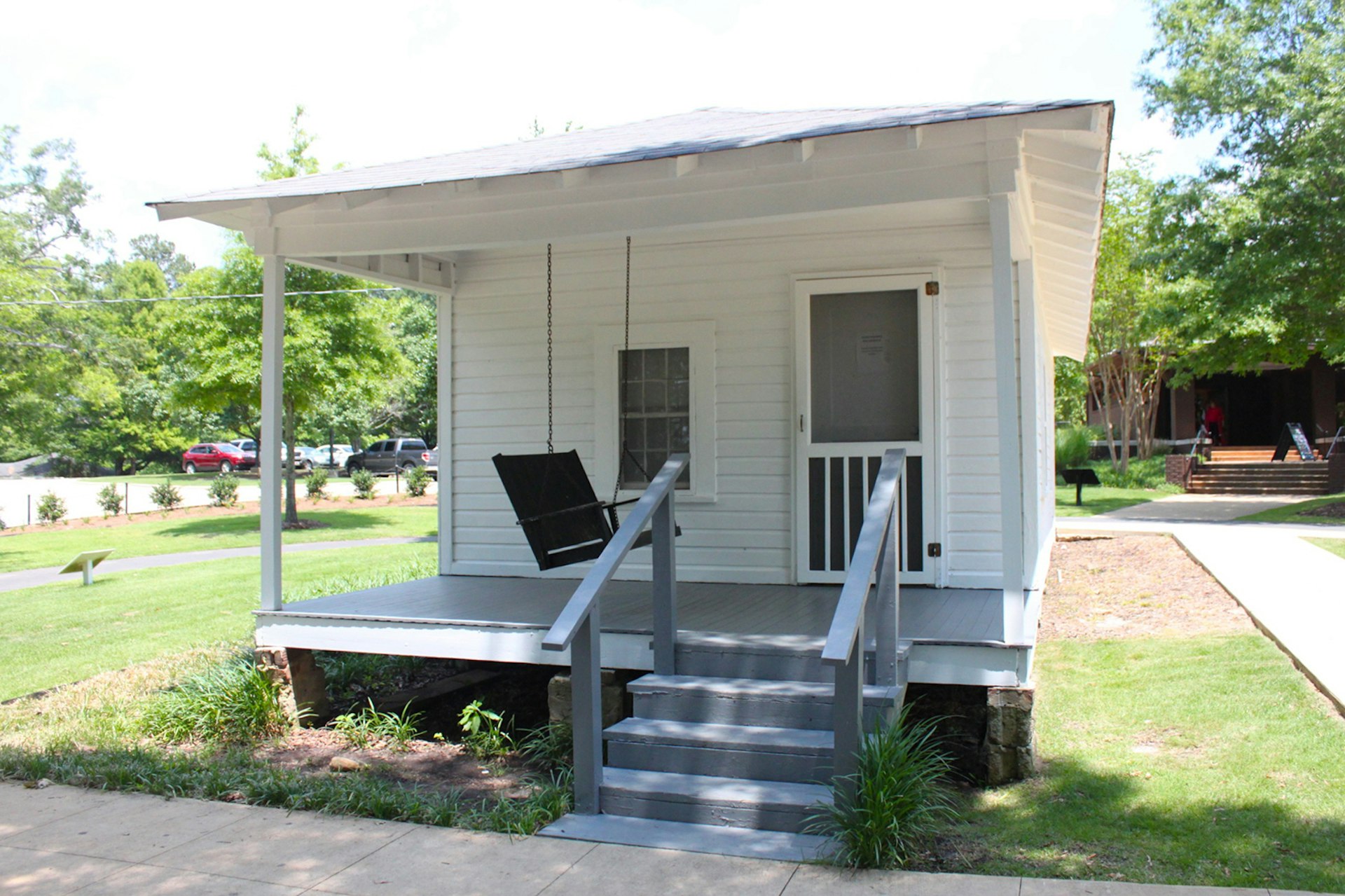 A small white shotgun shack with a front porch and a front porch swing sits on green grass under a blue sky in Tupelo, Mississippi