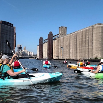 Several kayakers paddle past large concrete grain silos on the Buffalo River