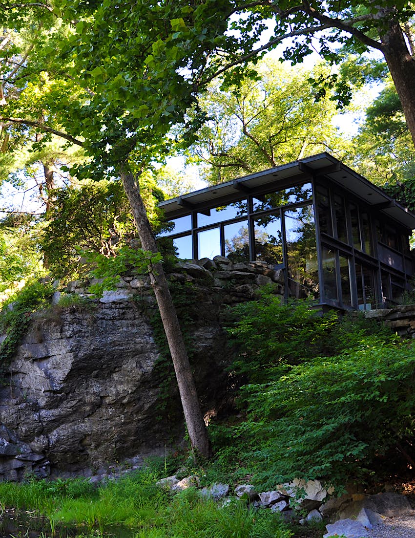 Exterior shot of Manitoga with glass front, sitting on rock face surrounded by trees and greenery Â© Manitoga