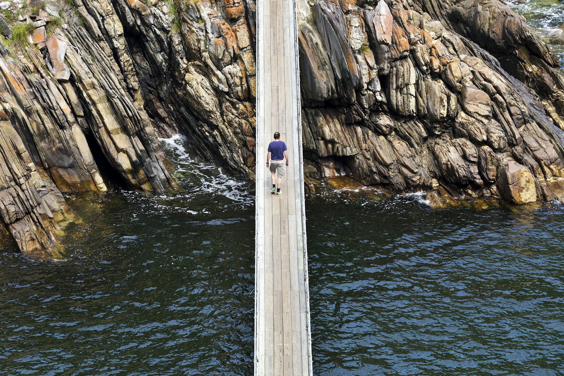 A man walking on wooden bridge across an inlet bound by striated rocky shores.