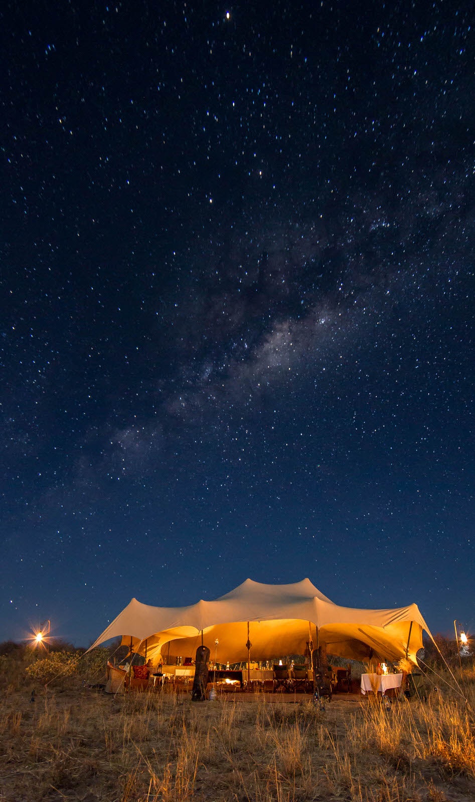 A stunning open-sided mess tent at a mobile safari camp, its canvas roof dramatically hung over a series of poles - sits lit by lanterns under a night sky full of stars © James Gifford / Lonely Planet