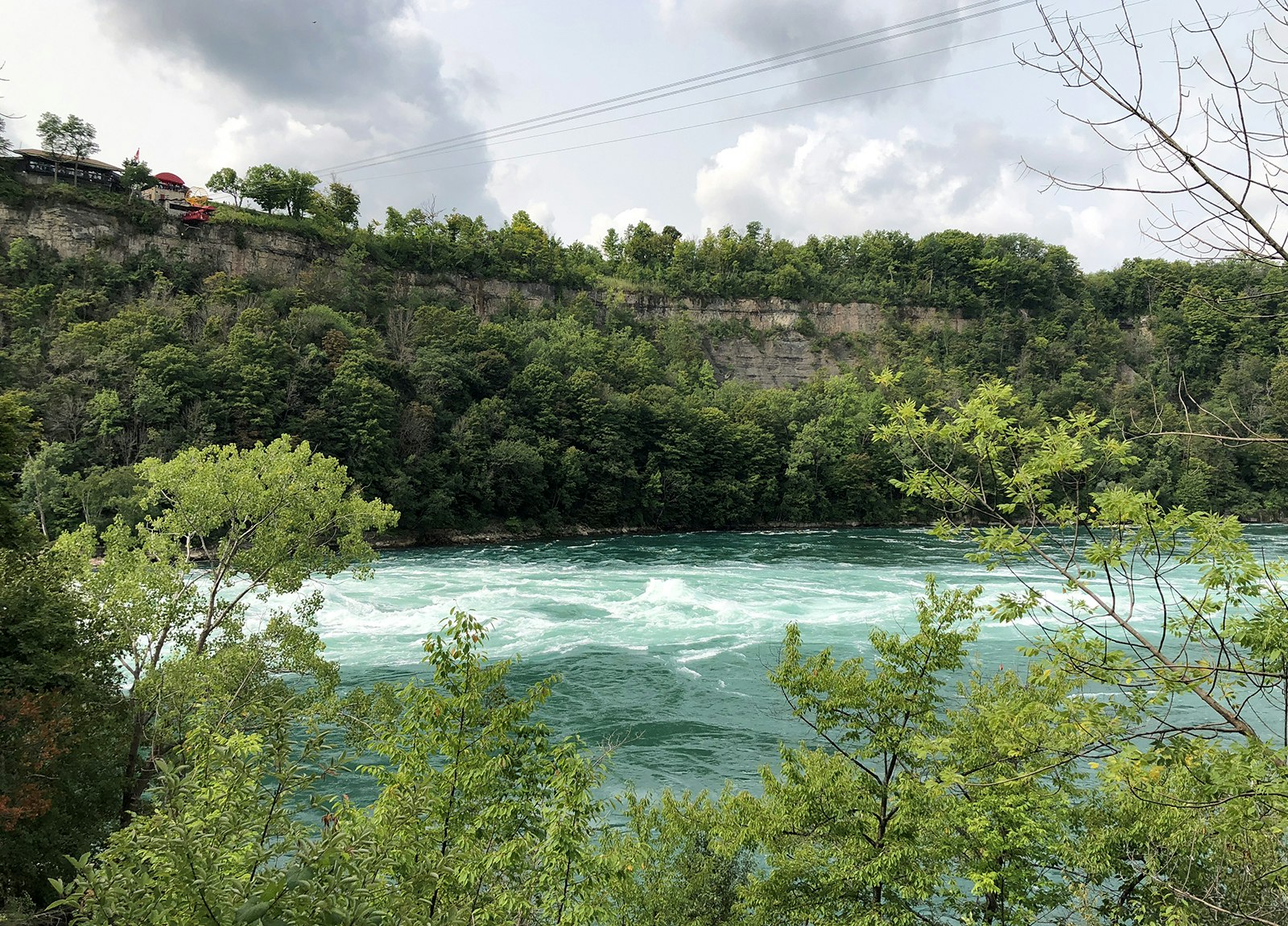 Vivid blue whirlpool with cliffs behind it and trees in front of it on a cloudy summer day in Niagara Gorge