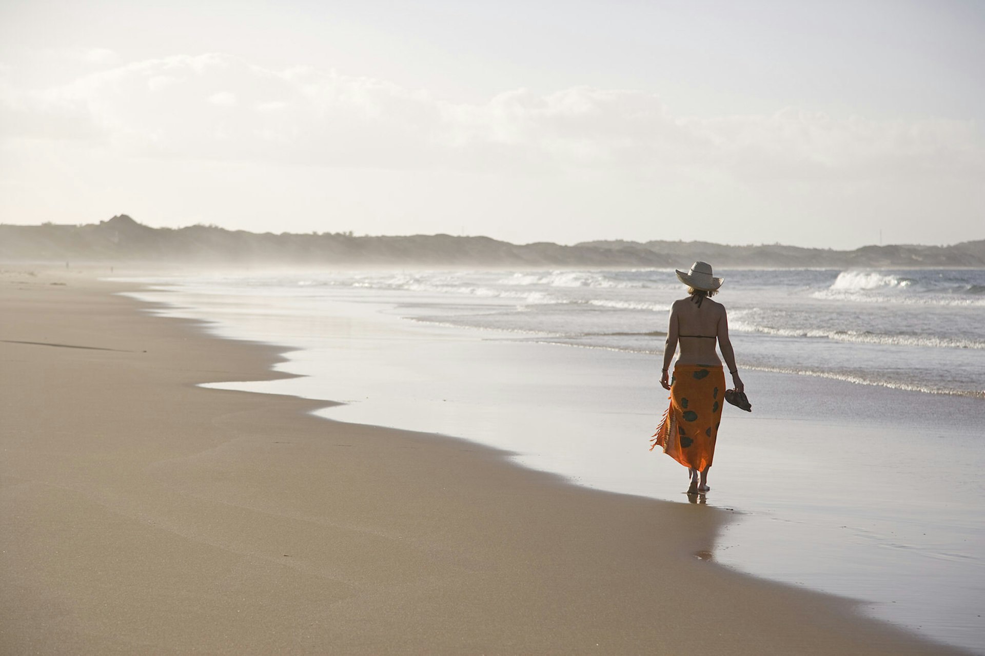 A women wearing a brimmed straw hat, sarong wrap and bikini top walks away from the camera along the wave strewn beach at Tofo © Julian Love / Getty Images