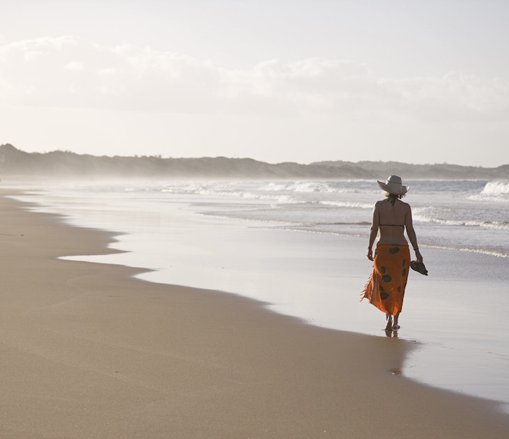 A women wearing a brimmed straw hat, sarong wrap and bikini top walks away from the camera along the wave strewn beach at Tofo © Julian Love / Getty Images