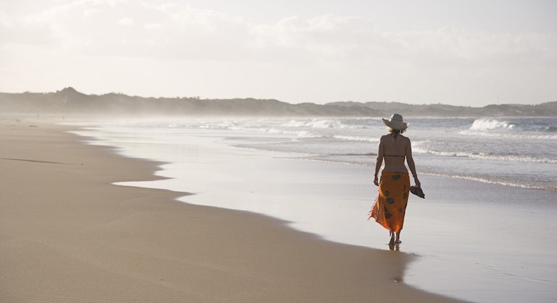 A women wearing a brimmed straw hat, sarong wrap and bikini top walks away from the camera along the wave strewn beach at Tofo © Julian Love / Getty Images
