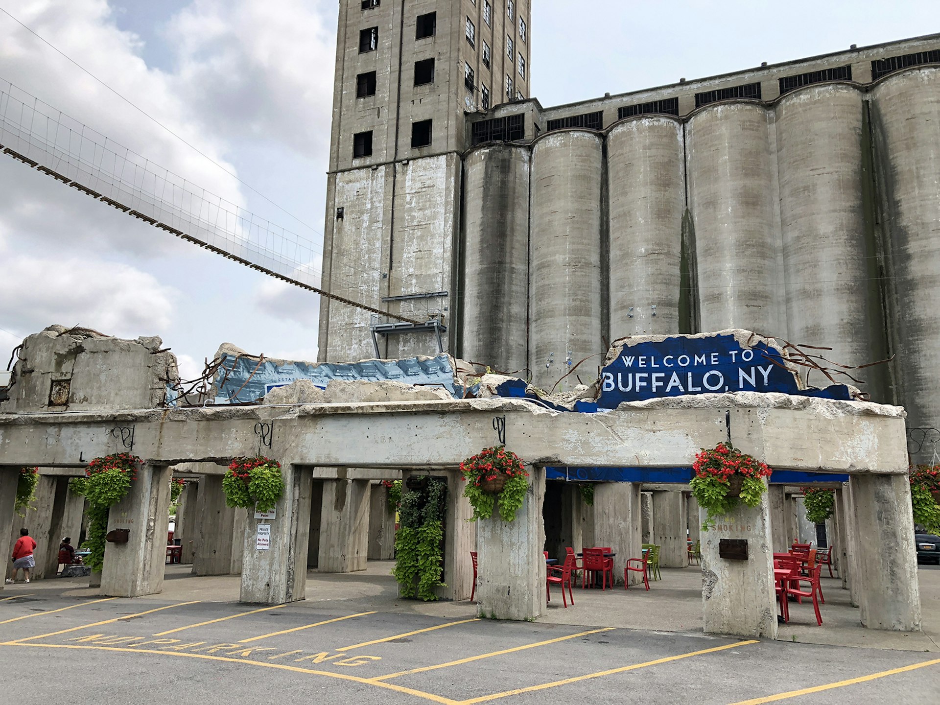 A rope bridge stretches between two grain silos on the zipline course at RiverWorks, above the broken foundations of two other grain silos topped with a painted 'Welcome to Buffalo' sign