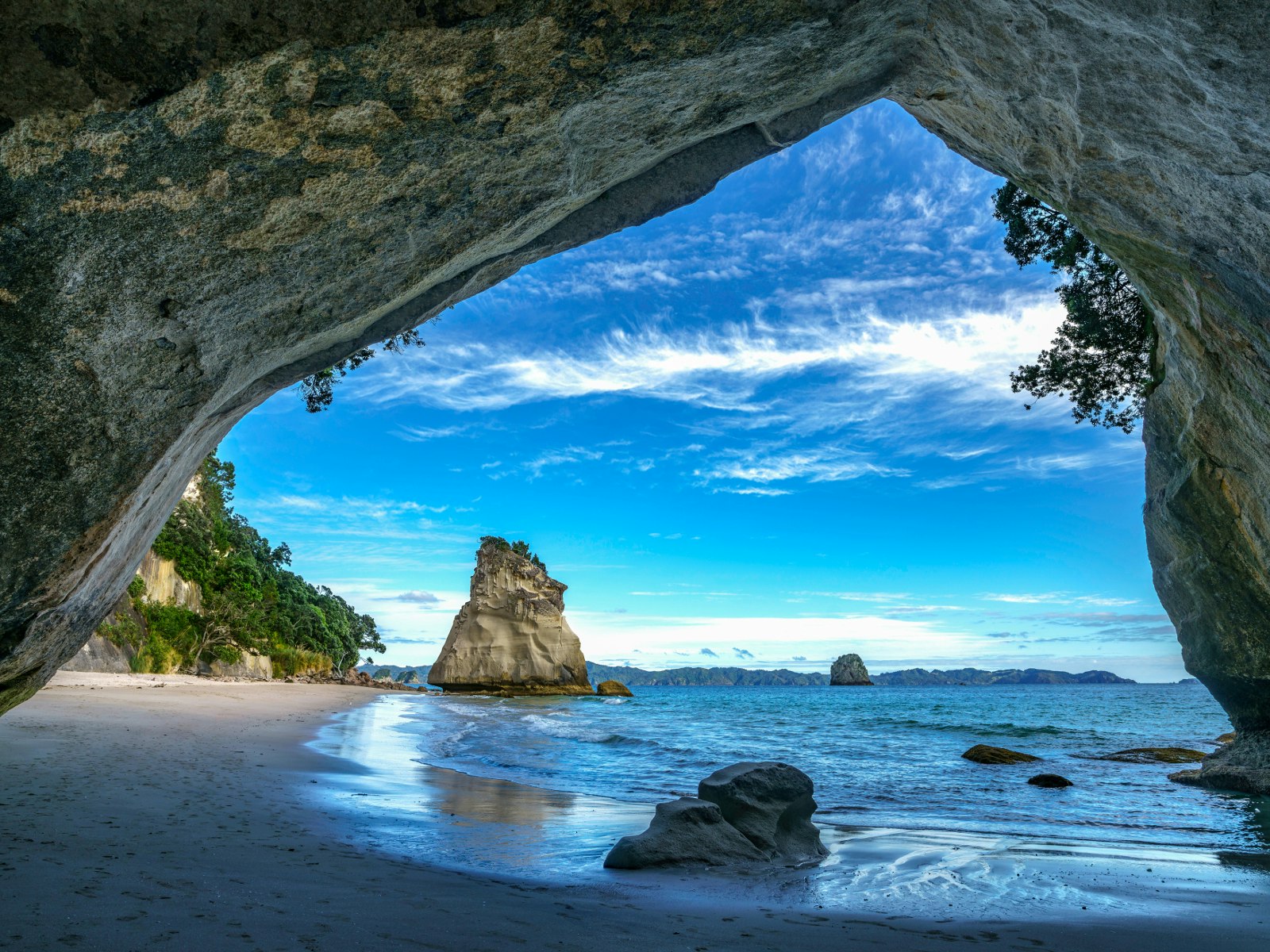 The shot is taken through a rock arch over Cathedral Cove beach, with a large freestanding rock formation at the end of the sweeping beach