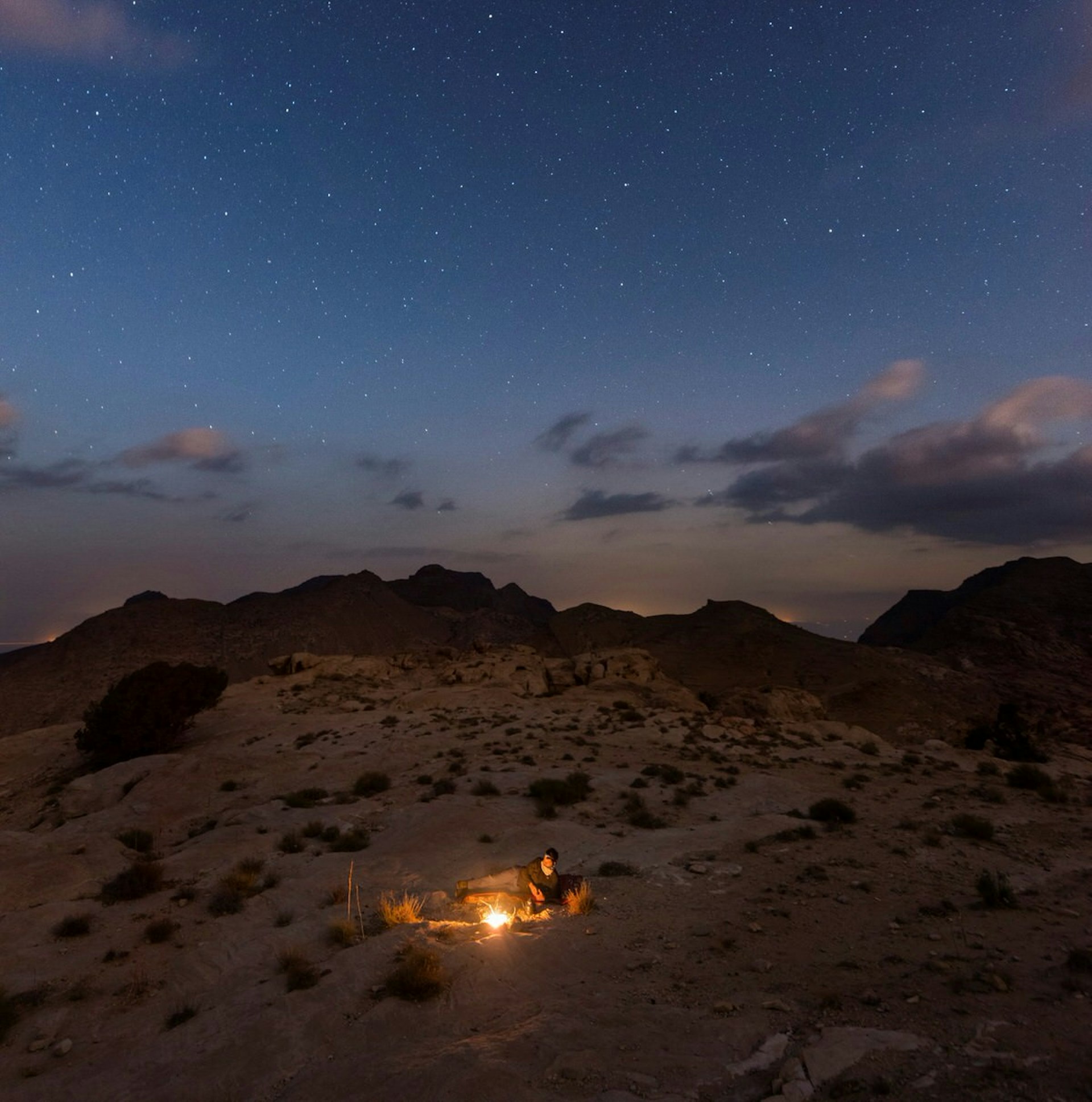 A hiker sits beside a lone fire with a star-filled sky above them 