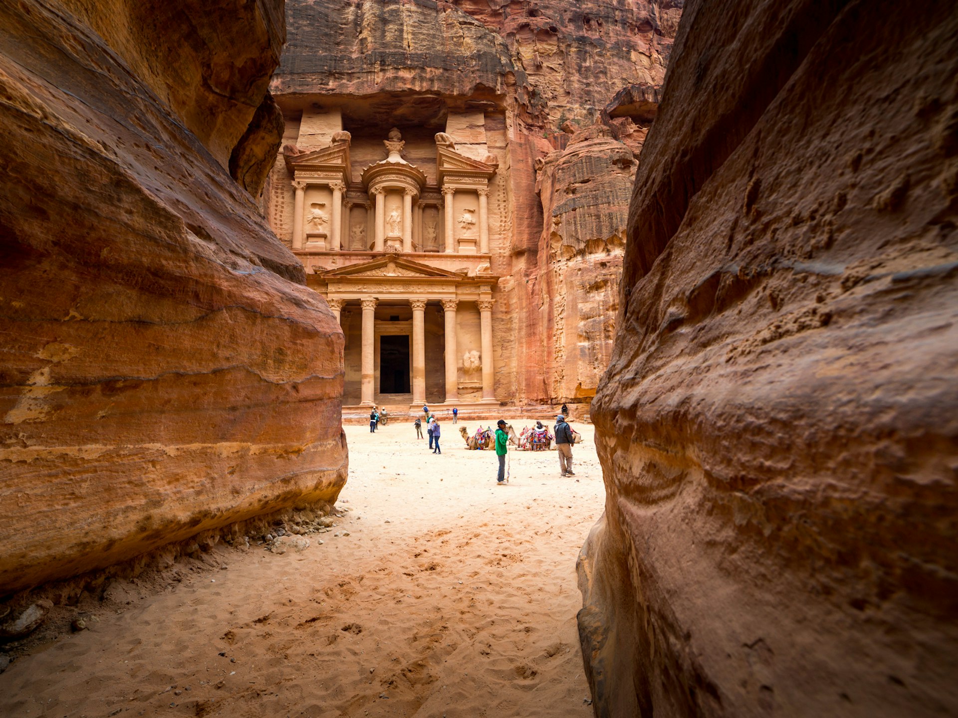 A glimpse of Petra's rock-hewn ancient city through a passageway in the rock. All the rock faces have a red hue, with the carvings themselves appearing more beige
