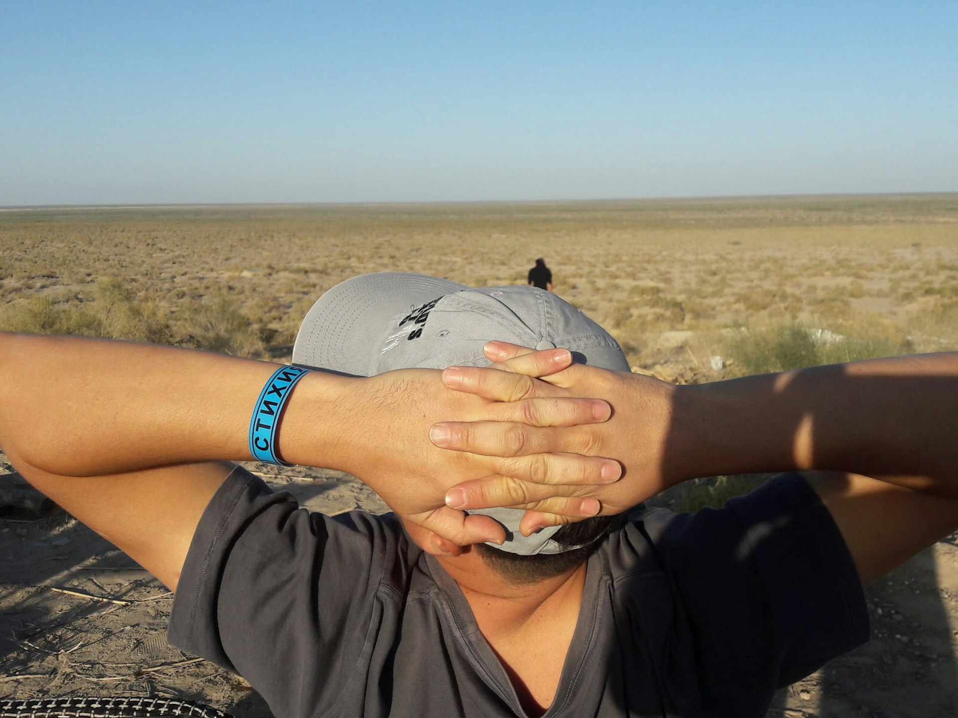 A man with his hands crossed behind his head is seen from behind, with the desert of the former Aral Sea in front of him