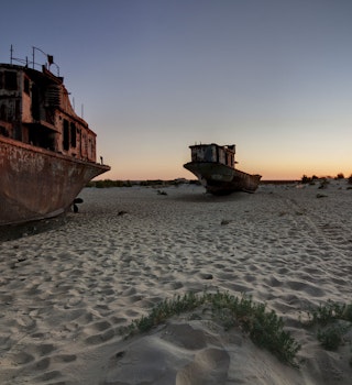 Rusting ships beached on sand with the sunset behind.