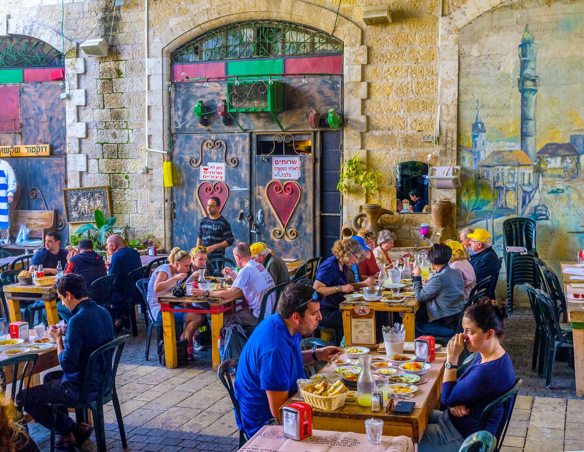 Patrons eat in an alley in Jaffa Flea Market, the wall lined with colourful murals and decorations.