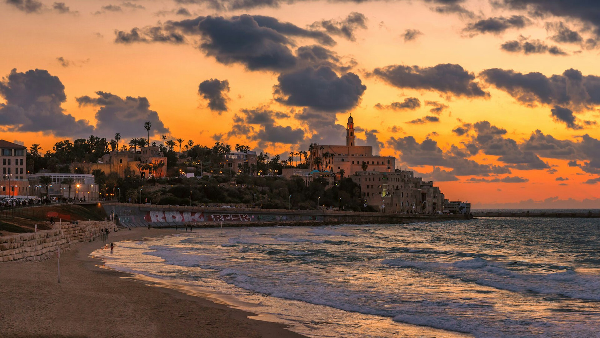 View from Jaffa, Tel Aviv, of a vivid orange sky as the sun sets over a headland covered in trees with a church at the far end.