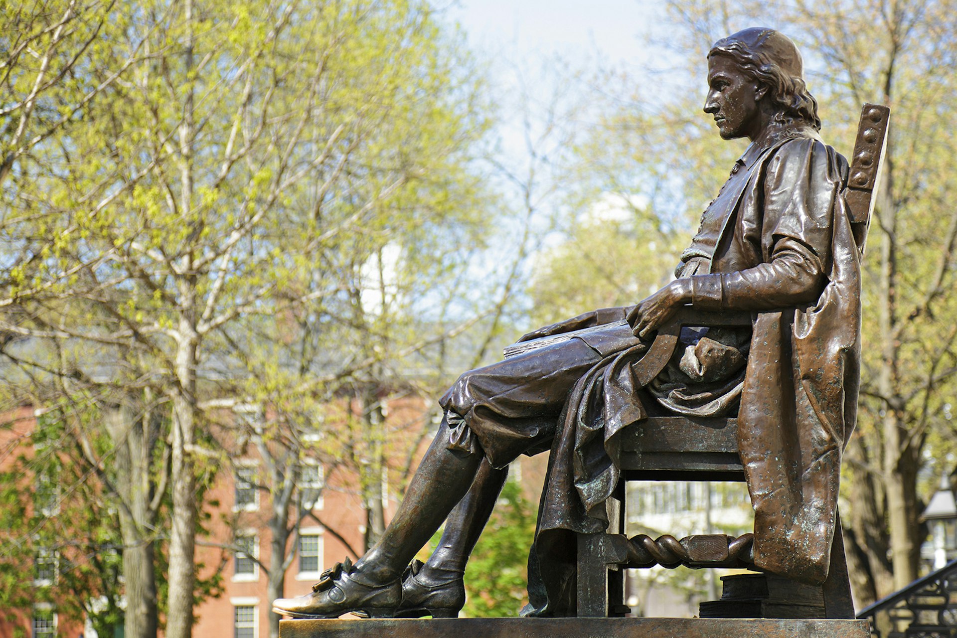 side view of bronze statue of a 17th century American man, John Harvard, with Harvard's red-brick buildings in the background