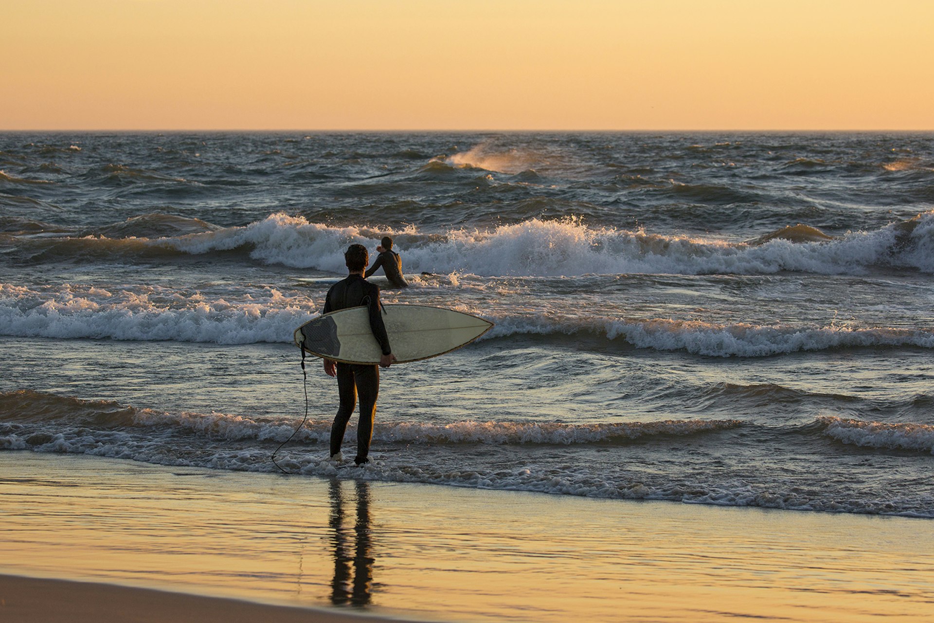 Man holding a surfboard and wearing a westuit stands on a white sand beach at sunset on Lake Michigan, watching a second man surf a wave