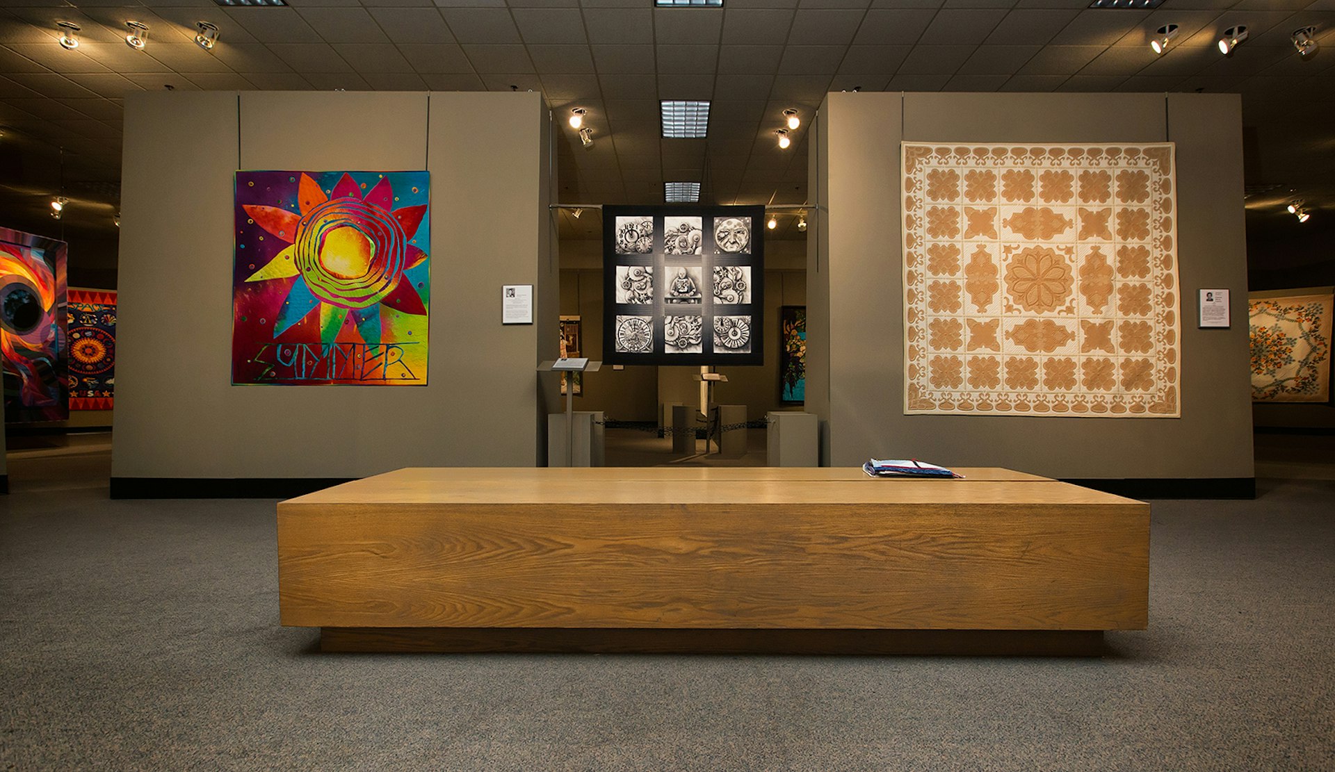 Wide shot of the gallery of the National Quilt Museum, with a modern, bright and abstract quilt hanging next to a traditional pattern in neutral colors