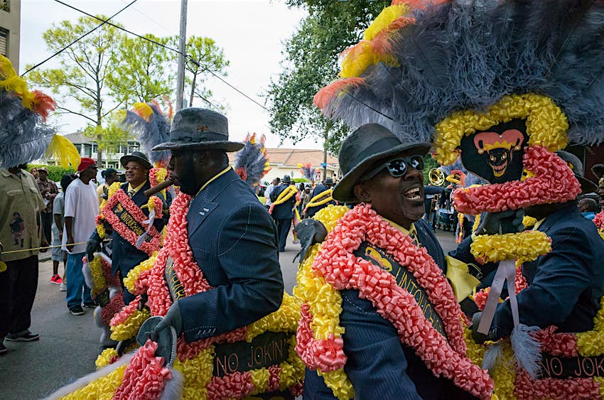 three men in black suits and yellow and pink garlands march in a second line in New Orleans on a cloudy day