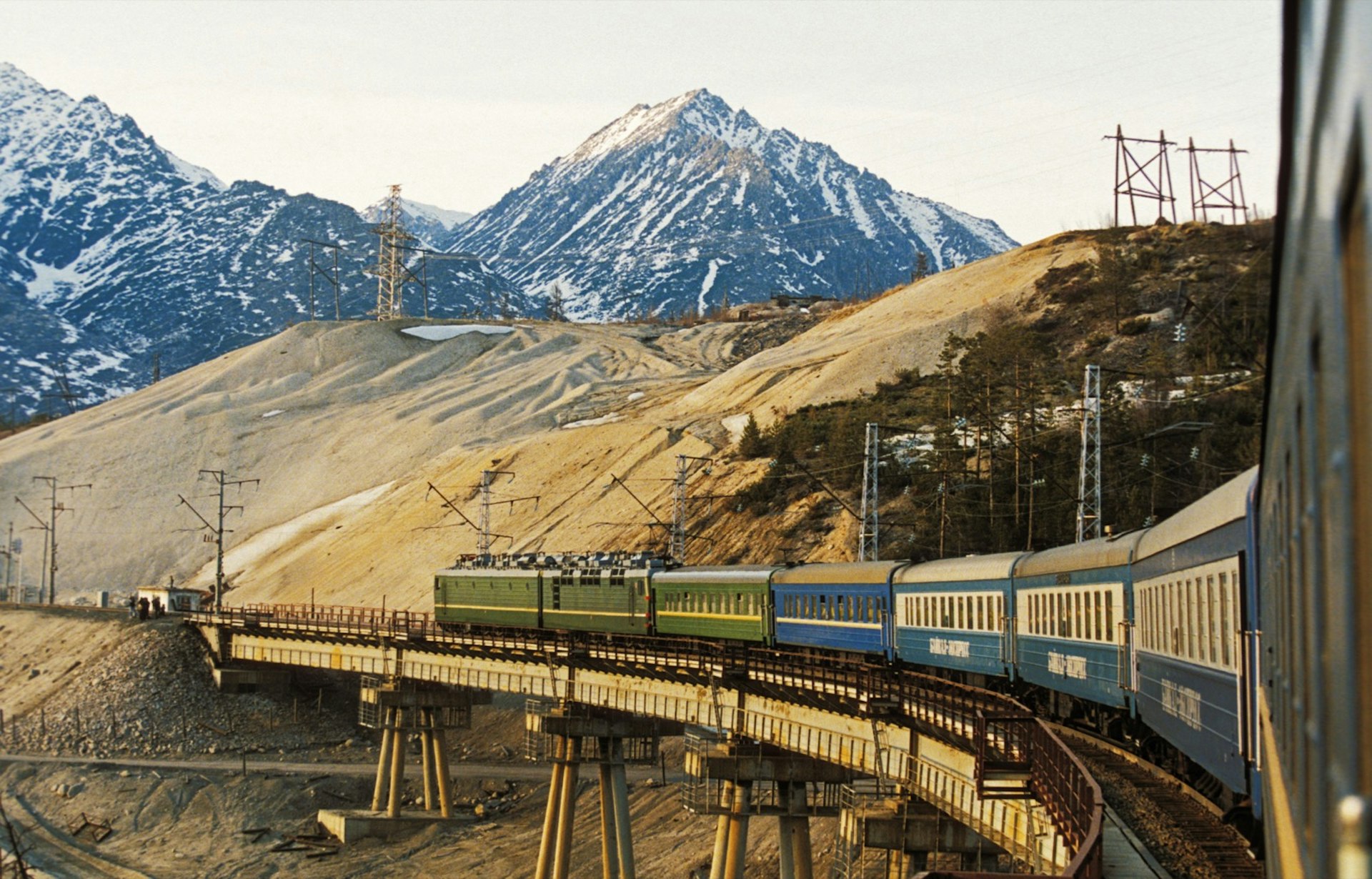 Dusty green and blue train cars rumble over a curved bridge at the base of a snow-capped mountain in Russia; amazing train journeys