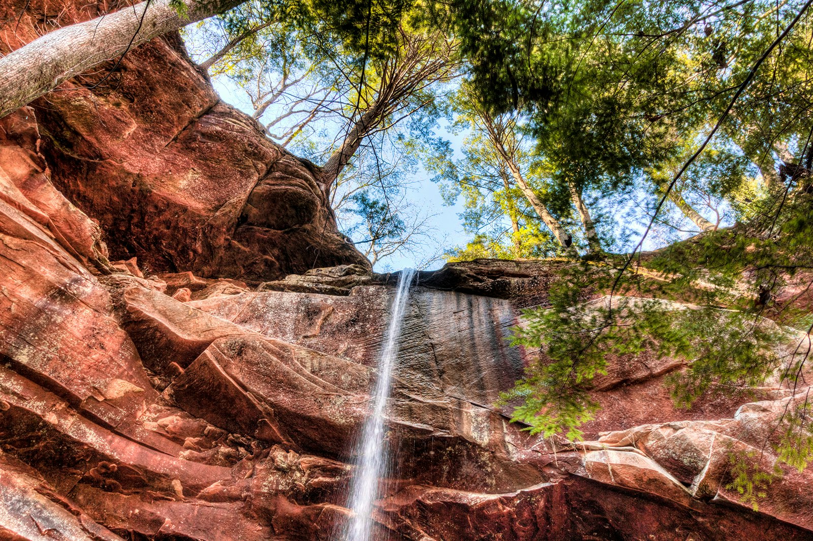 Upward shot of a waterfall trickling over the red rock walls of a canyon, with blue sky and green trees overhead