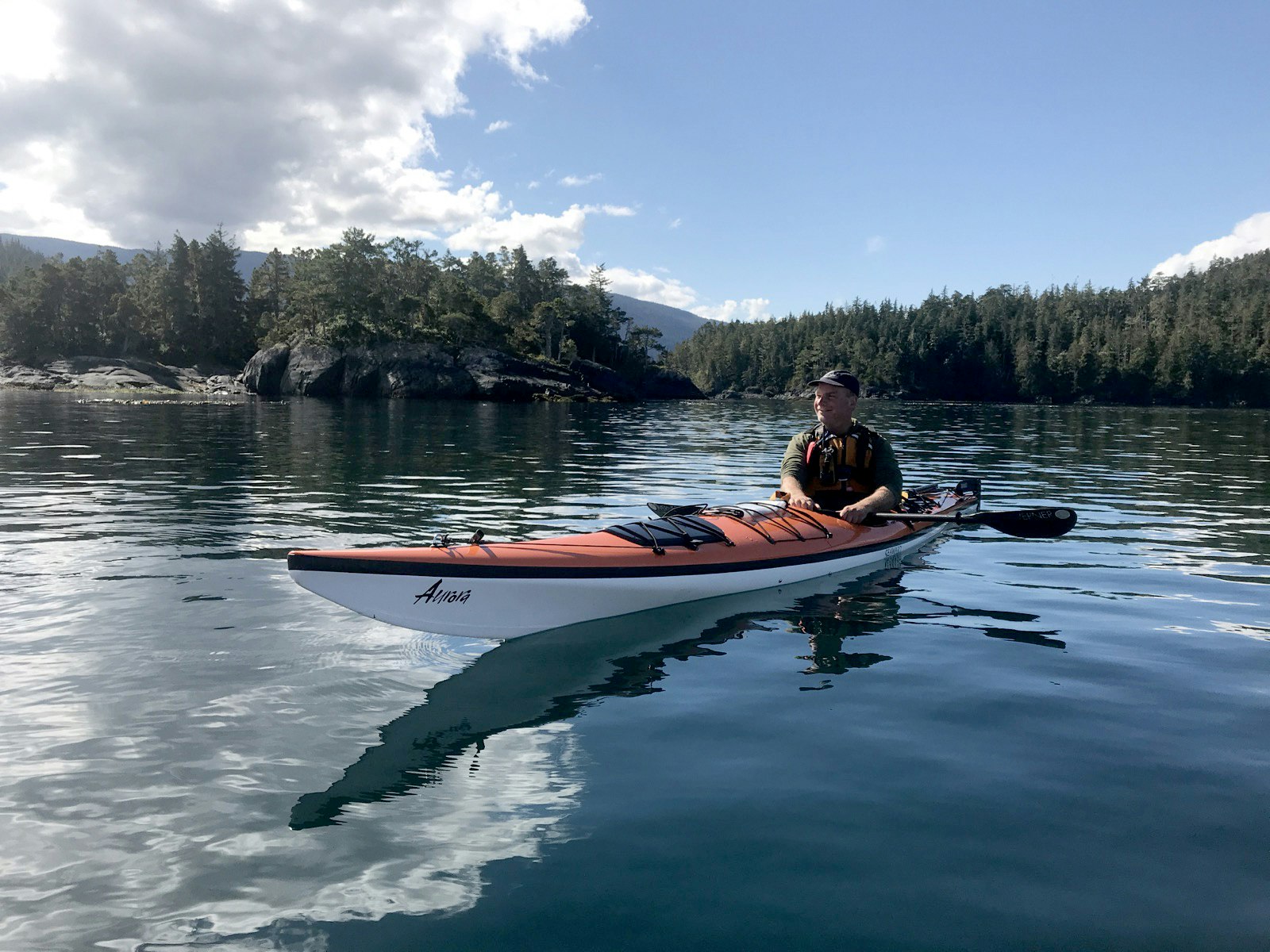 A man paddles an orange and white kayak across a very still inlet in British Columbia.