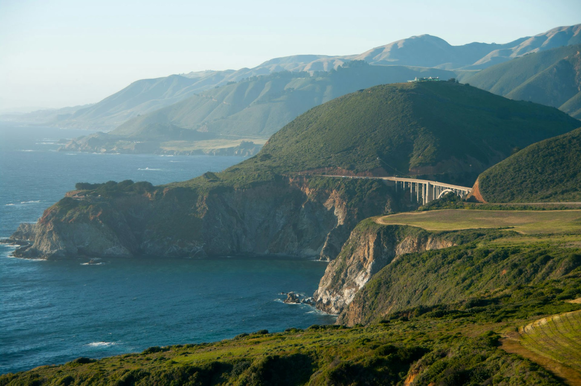 Rolling hills end in cliffs that border the ocean, a single span bridge stretches across the hills. 