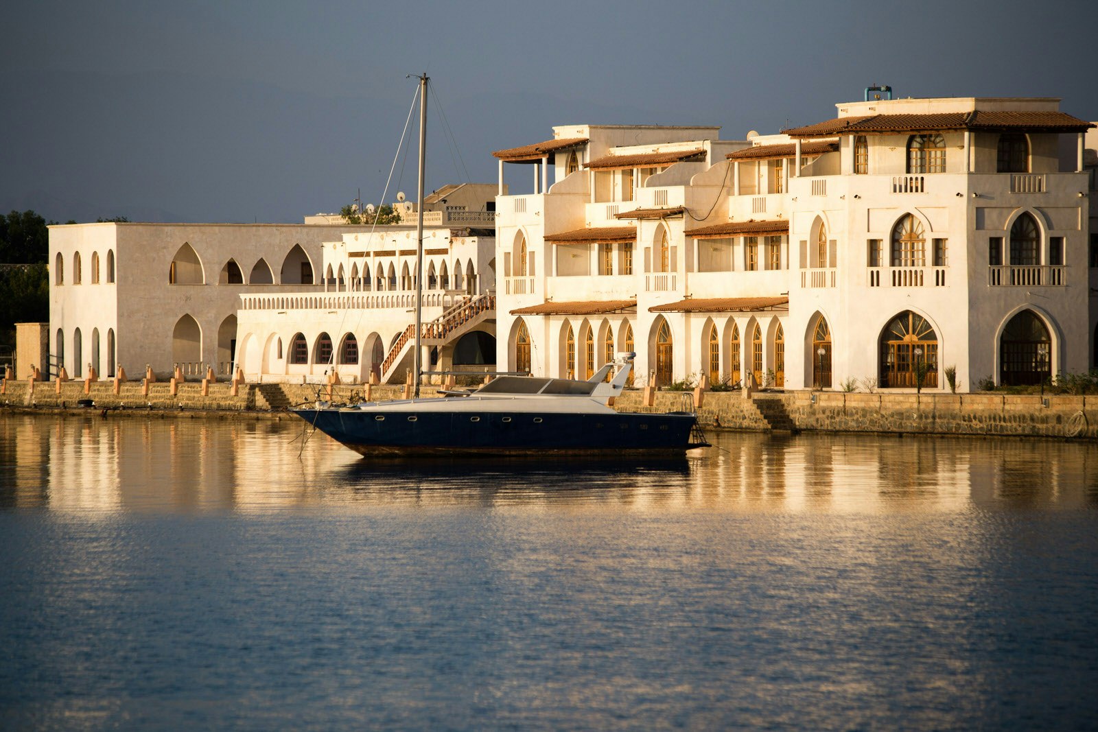 Warm light cast by a setting sun lights a row of three storey, sand-coloured colonial buildings from the Italian era in Massawa, Eritrea - in the foreground are flat waters and a modern powerboat at anchor