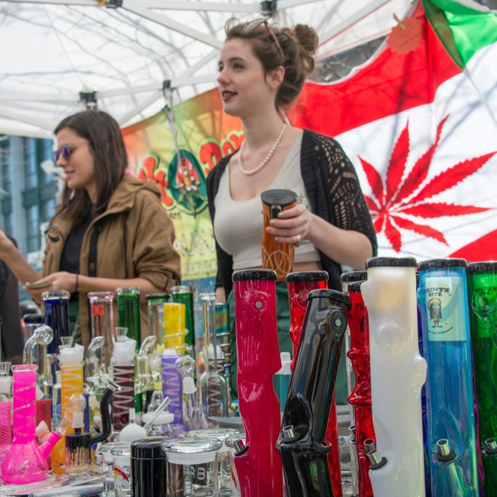 Two women sell bongs and other marijuana items at an outdoor stall, with a flag of Canada in the background that has the maple leaf replaced with a marijuana leaf.