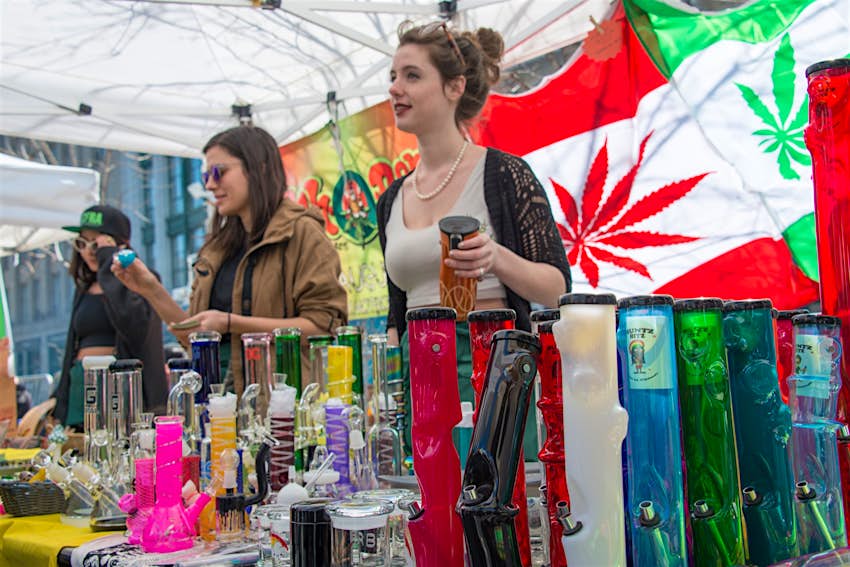 Features - Toronto 420 Rally for Marihuana in Dundas Square Two women sell bongs and other marijuana items at an outdoor stall, with a flag of Canada in the background that has the maple leaf replaced with a marijuana leaf.