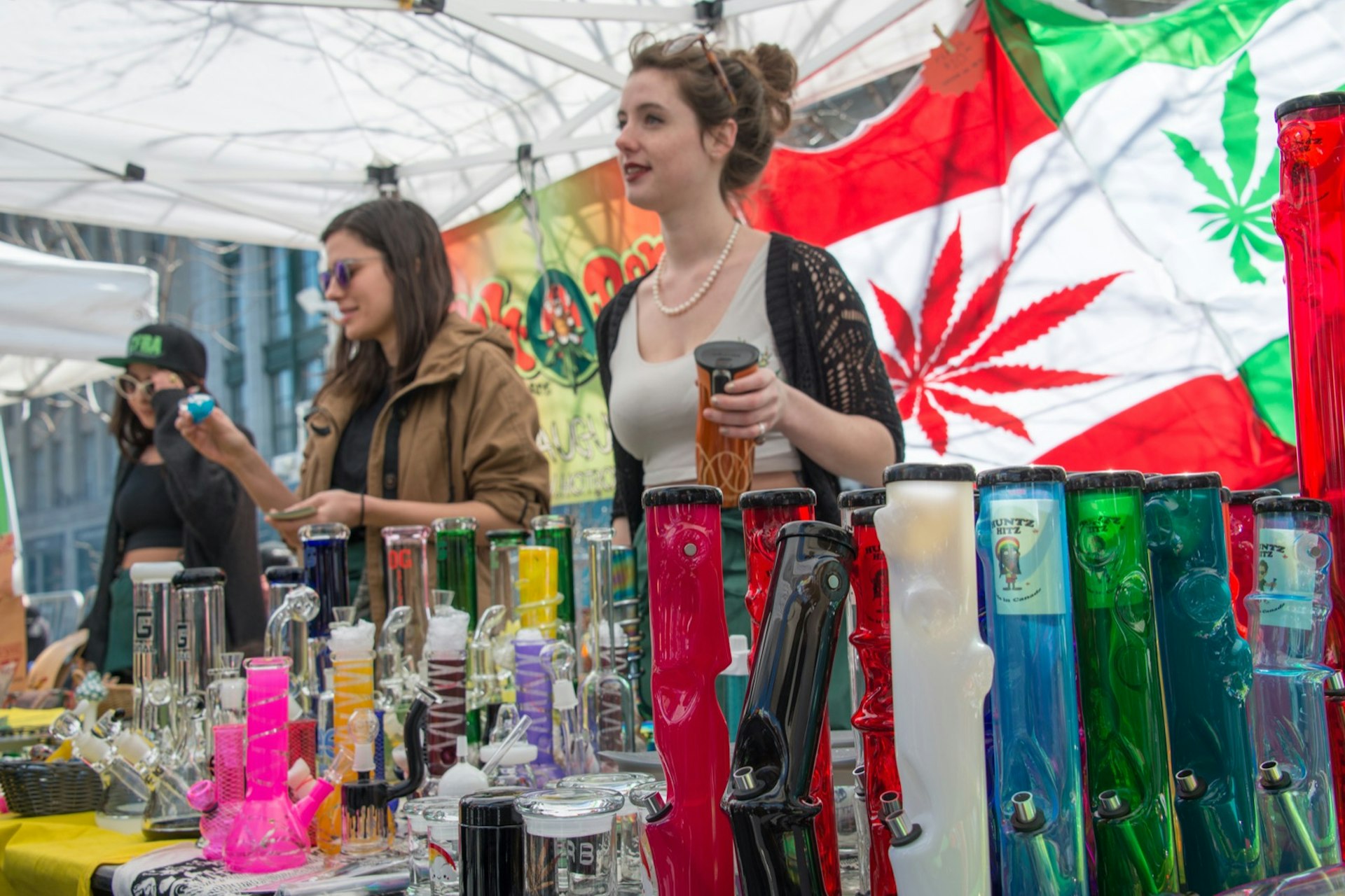 Two women sell bongs and other marijuana items at an outdoor stall, with a flag of Canada in the background that has the maple leaf replaced with a marijuana leaf.