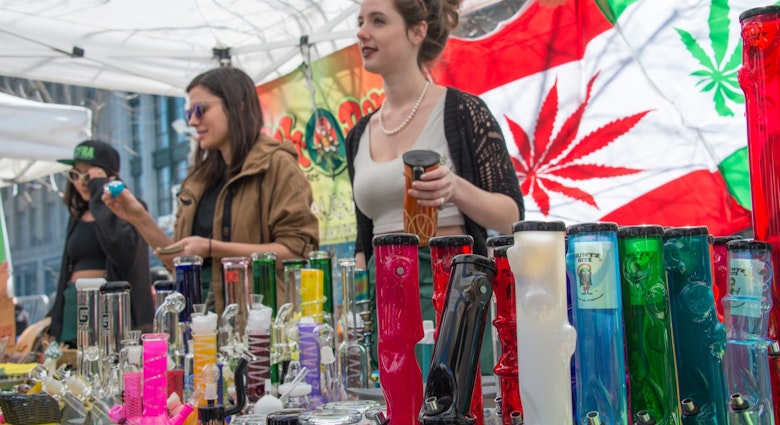 Two women sell bongs and other marijuana items at an outdoor stall, with a flag of Canada in the background that has the maple leaf replaced with a marijuana leaf.