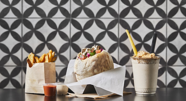 Fries in a paper cup, a falafel in a wax-paper wrapper and a frosty milkshake with a yellow straw sit on a counter in front of a black-and-white tile background at Goldie's in Philadelphia