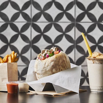 Fries in a paper cup, a falafel in a wax-paper wrapper and a frosty milkshake with a yellow straw sit on a counter in front of a black-and-white tile background at Goldie's in Philadelphia